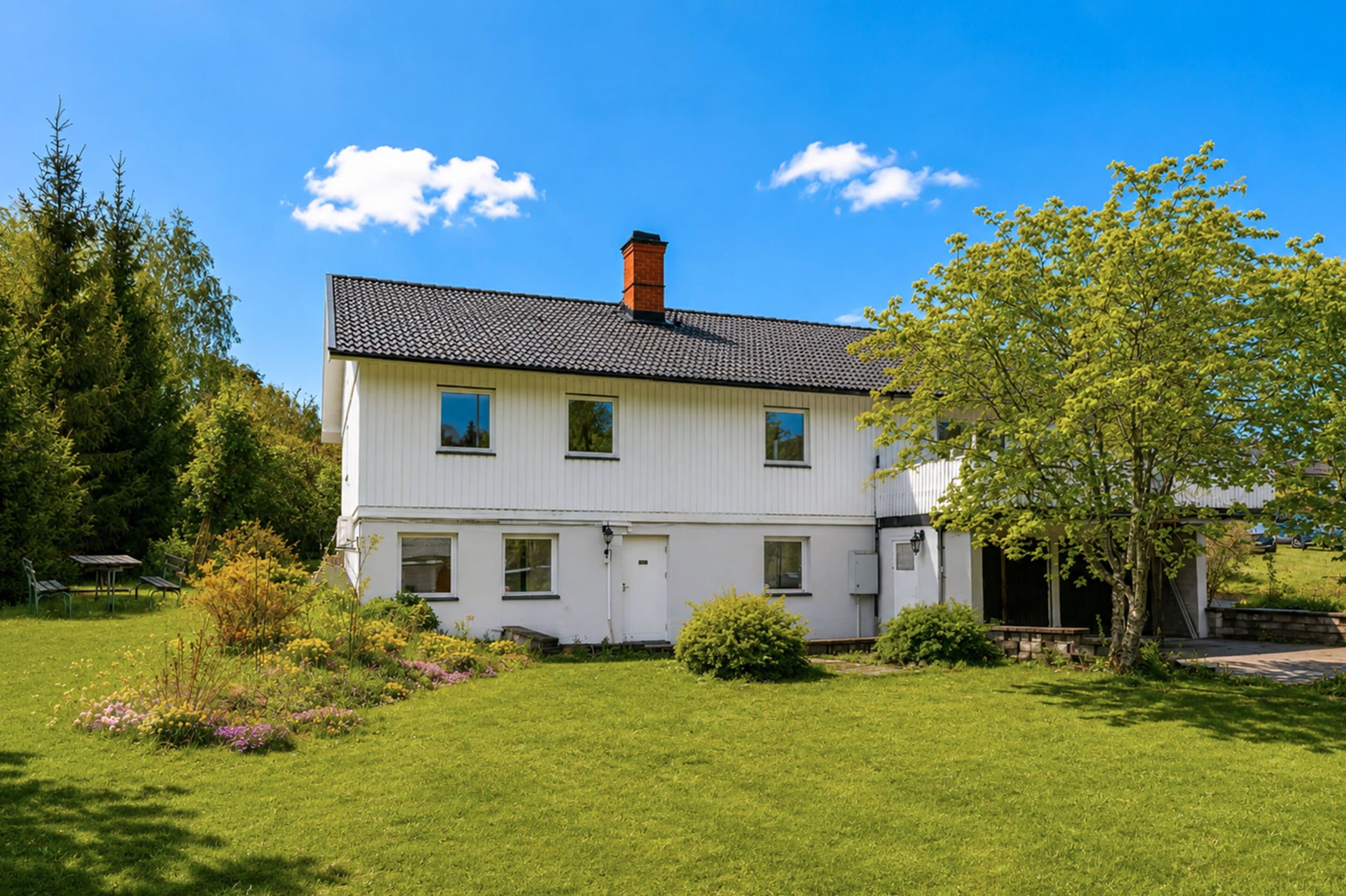 Vacker vita villa med svart tak och röd skorsten, omgiven av grön trädgård och blomsterrabatter. Lugnt läge bland träd med utsikt över gräsmark.